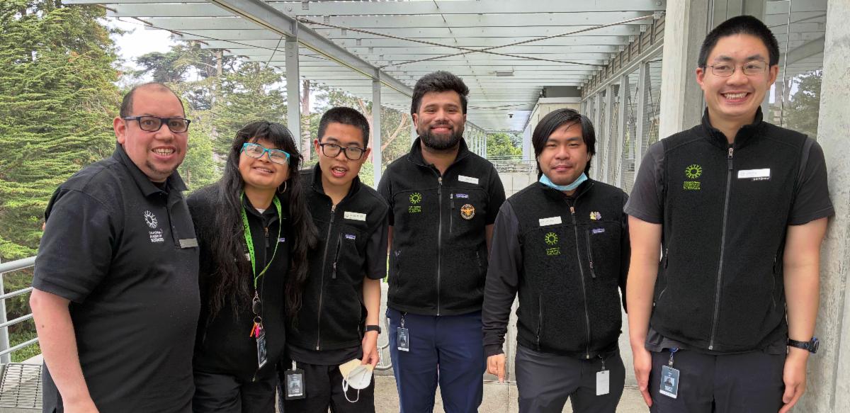 Group photo of recent Cal Academy interns in matching dark staff shirts with name badges, standing together on a covered outdoor walkway with trees and buildings in the background.