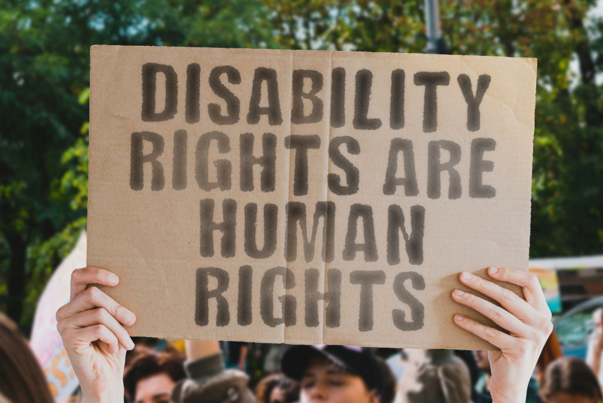 A crowd holds up a large sign that reads “DISABILITY RIGHTS ARE HUMAN RIGHTS” outdoors, with trees and other people visible in the background.