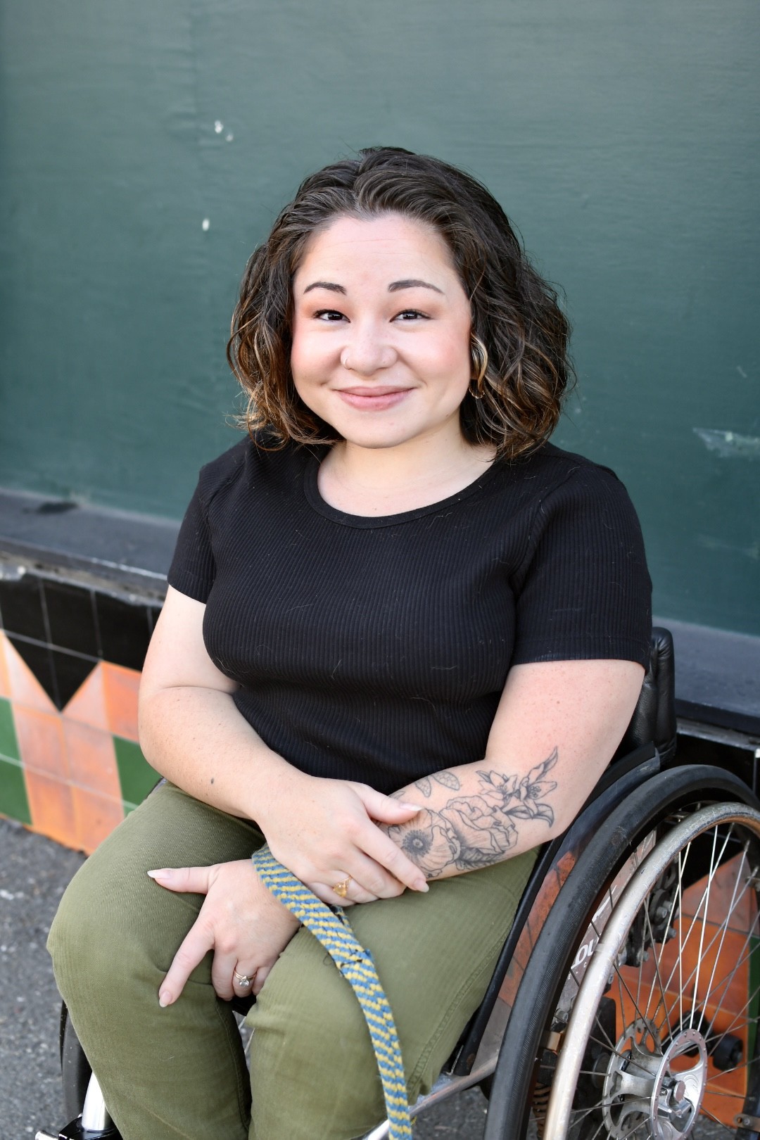 Rosemary with dark, curly hair and a black shirt smiles at the camera, seated in a wheelchair with a tattoo on their arm.