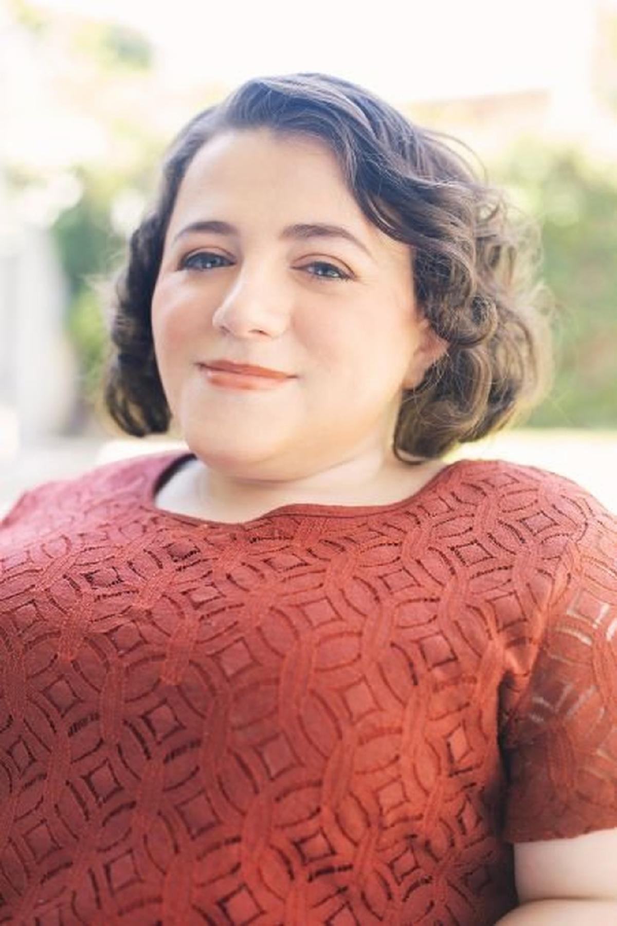 Kristen Lopez with dark, curly hair wearing a maroon lace top rests their arms on a table.