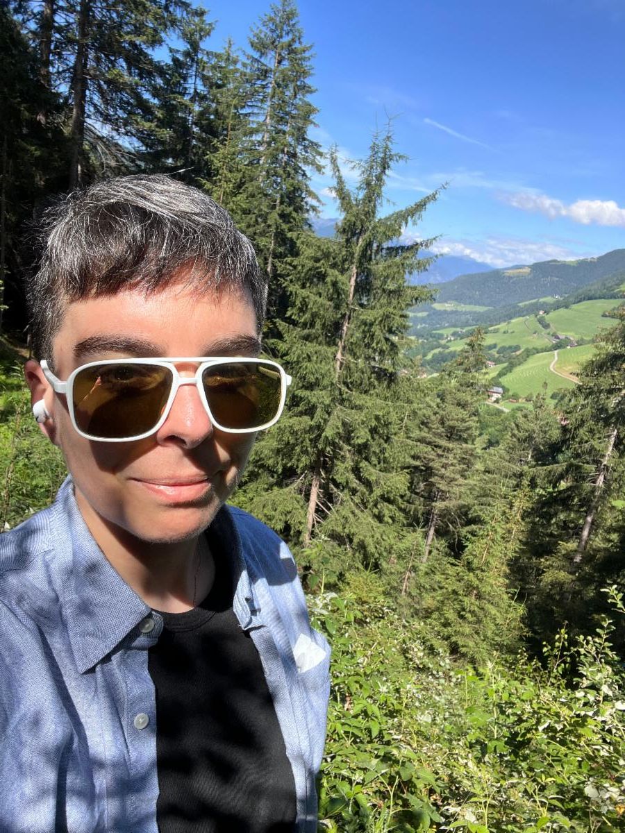 A selfie of Risa Shoup. A white person with short black and white hair stands smiling on a mountainside. Pine trees tower into a blue sky.