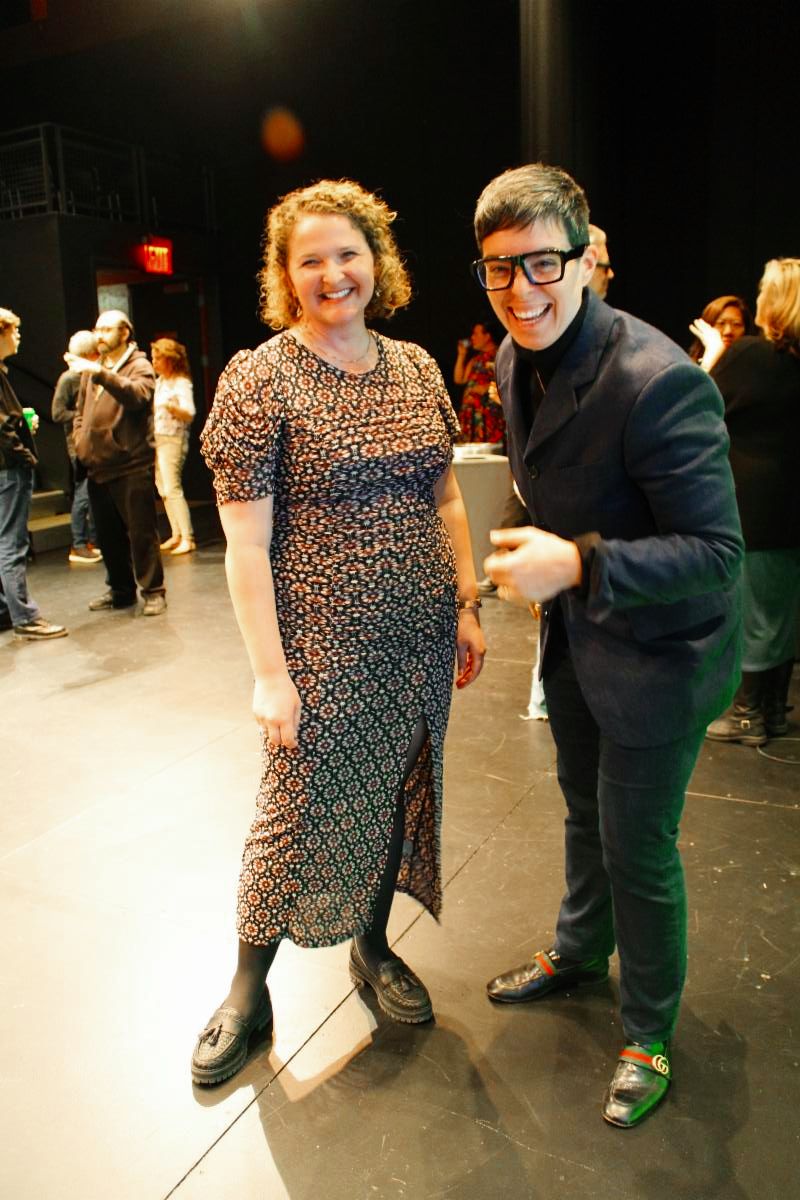 Risa Shoup and Talia Corren stand together at an ART New York event. Risa wears a black suit and thick black framed glasses and laughs. Talia wears a long flowery dress. 