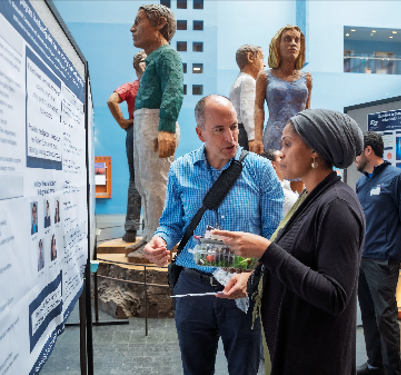 Photo from HEAR Symposium 2025 with a poster presenter talking about her research to an attendee.