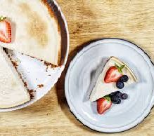 Photo illustration of cheesecake with one slice cut out on left and piece of cheesecake on plate to the right on a wooden table top.