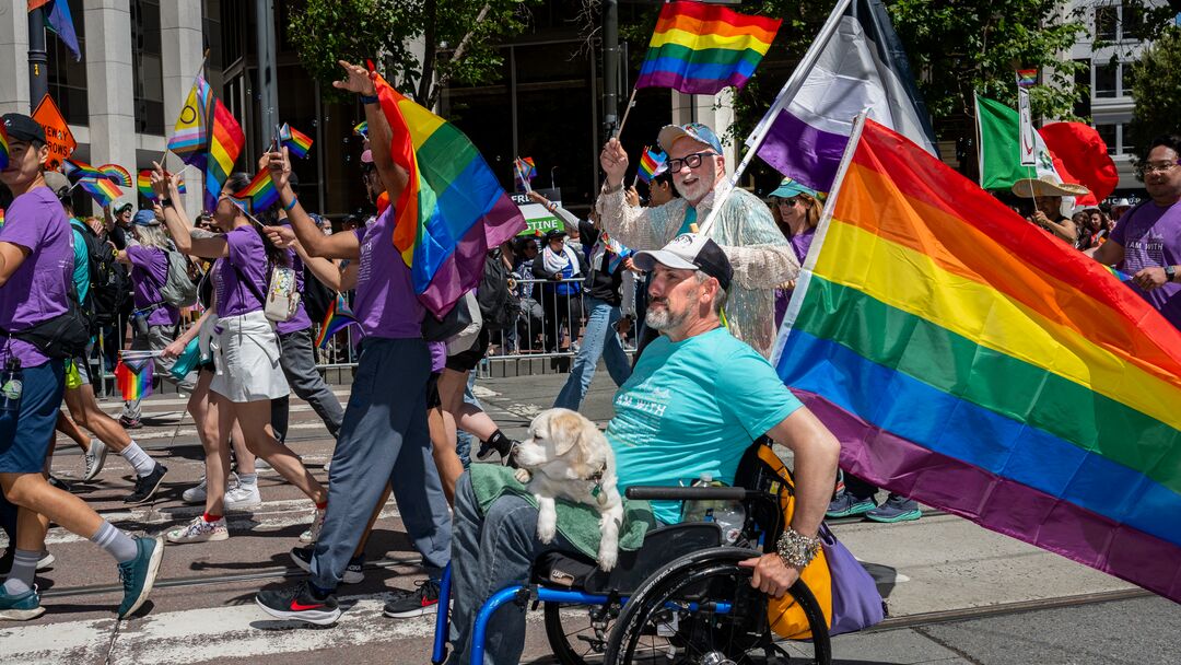 Photo from UCSF in Pride parade in 2024 with person with grey beard moving through parade with wheelchair and dog on his lap. A person behind him smiles and rainbow flags are seen. The mood is joyous.