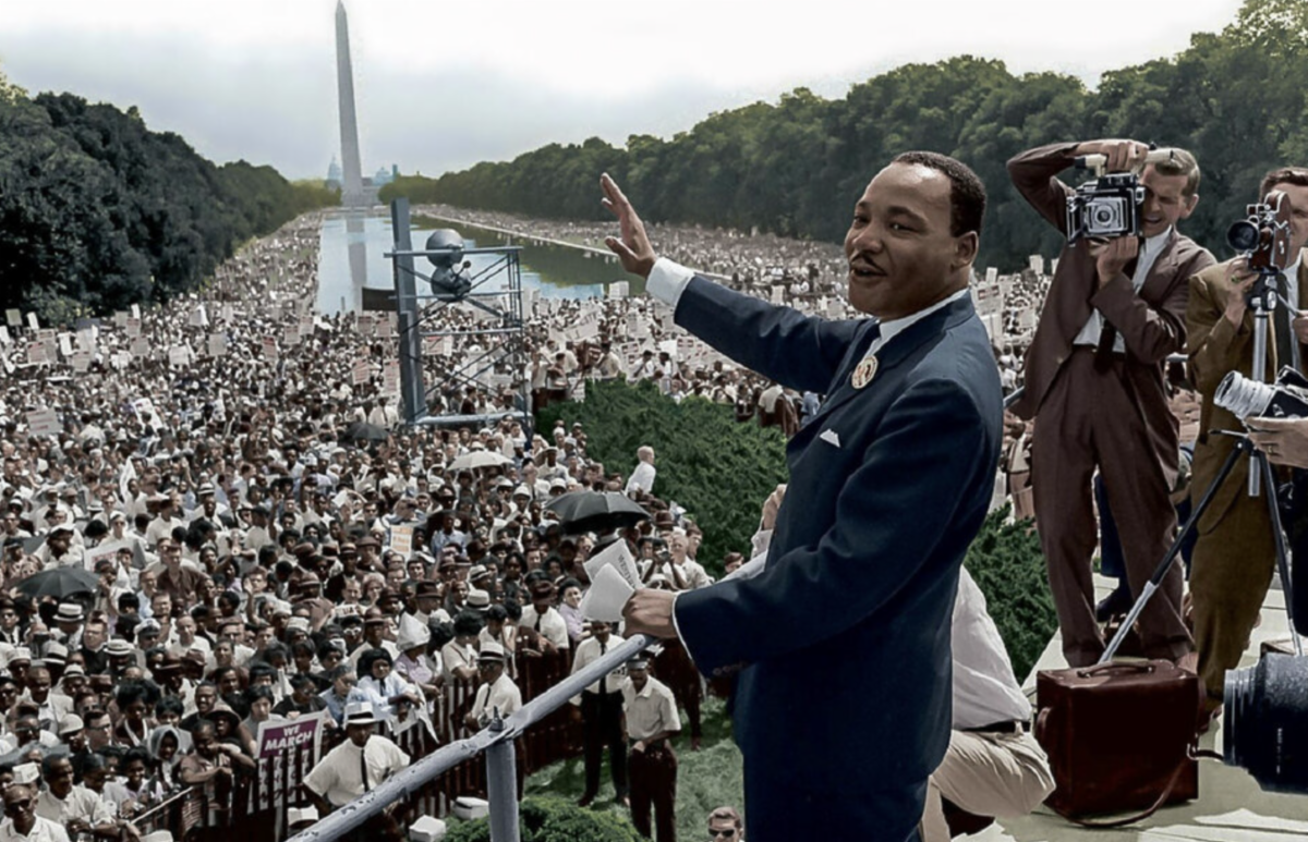 Photo of Dr. Martin Luther King, Jr., dressed in suit, waving to historically large crowd gathered for the march. Photographers are at right of photo taking photos of Dr. King. 