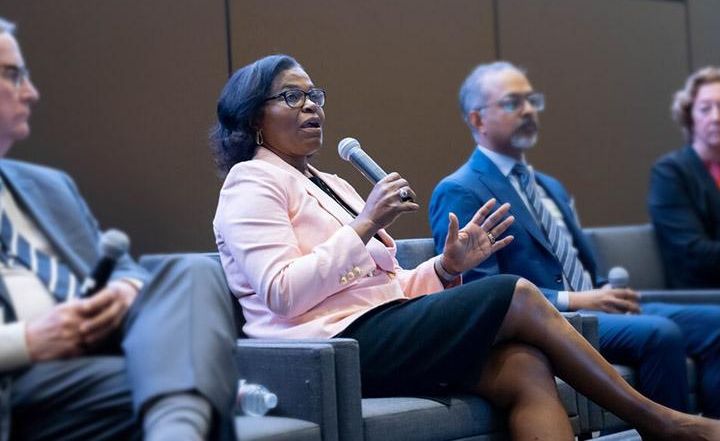 Photo of panelists sitting in row at symposium, indoors, with central speaker, Renee Navarro, at center, holding a microphone.