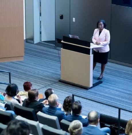 Photo of Vice Chancellor Renee Navarro speaking at podium with pink blazer on standing in front of screen with audience listening