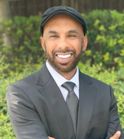 Photo of Mairaj Syed smiling wearing grey cap with grey blazer and white shirt. He is outside and smiling.