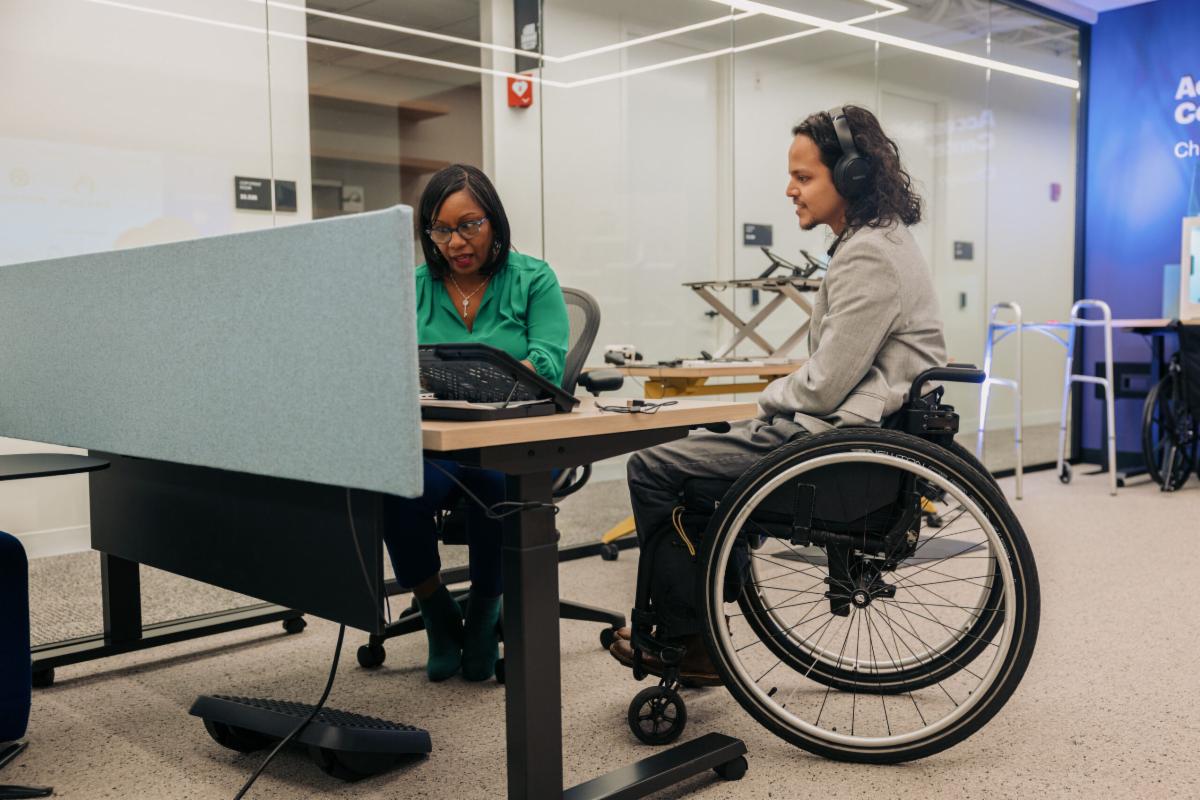 Two colleagues work together in an accessibility solutions center. One is wearing noise cancelling headphones and uses a wheelchair. The other reviews items with him on a laptop
