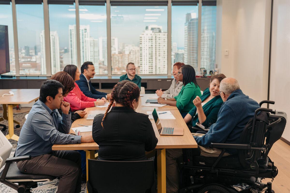 A group of diverse professional representing various disability identities gather in a meeting around a conference table. Windows overlook a city skyline.