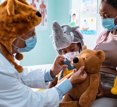 A healthcare provider wears a teddy bear hat and shows a young patient how to use an inhaler with a teddy bear.