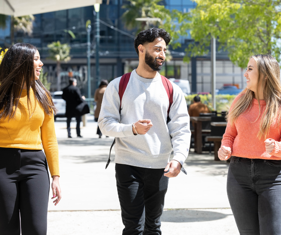Photo of three people walking outside, smiling and talking, on a sunny day. 