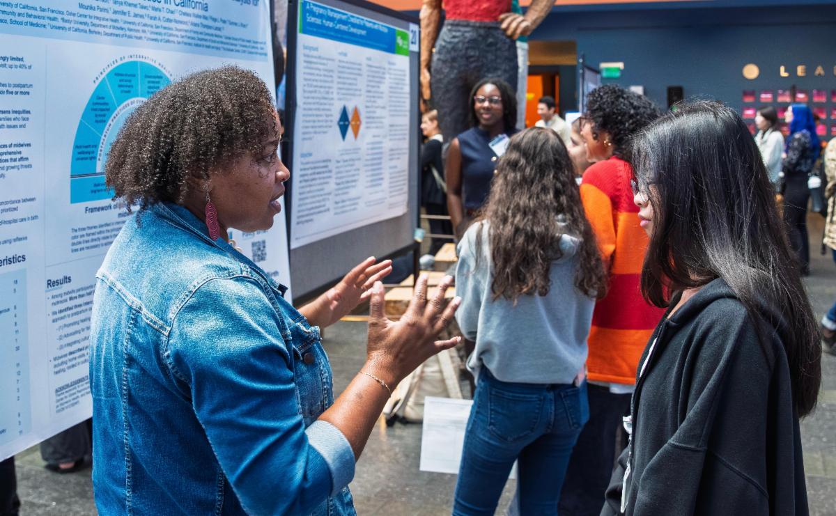 Photo of HEAR Symposium presenter talking to attendee indoors in Mission Bay Conference Center with posters seen in background. 