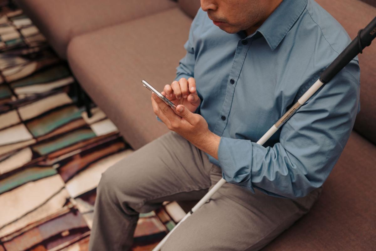 A man who is blind sits in an office as he navigates his phone. His white cane rests against his body.