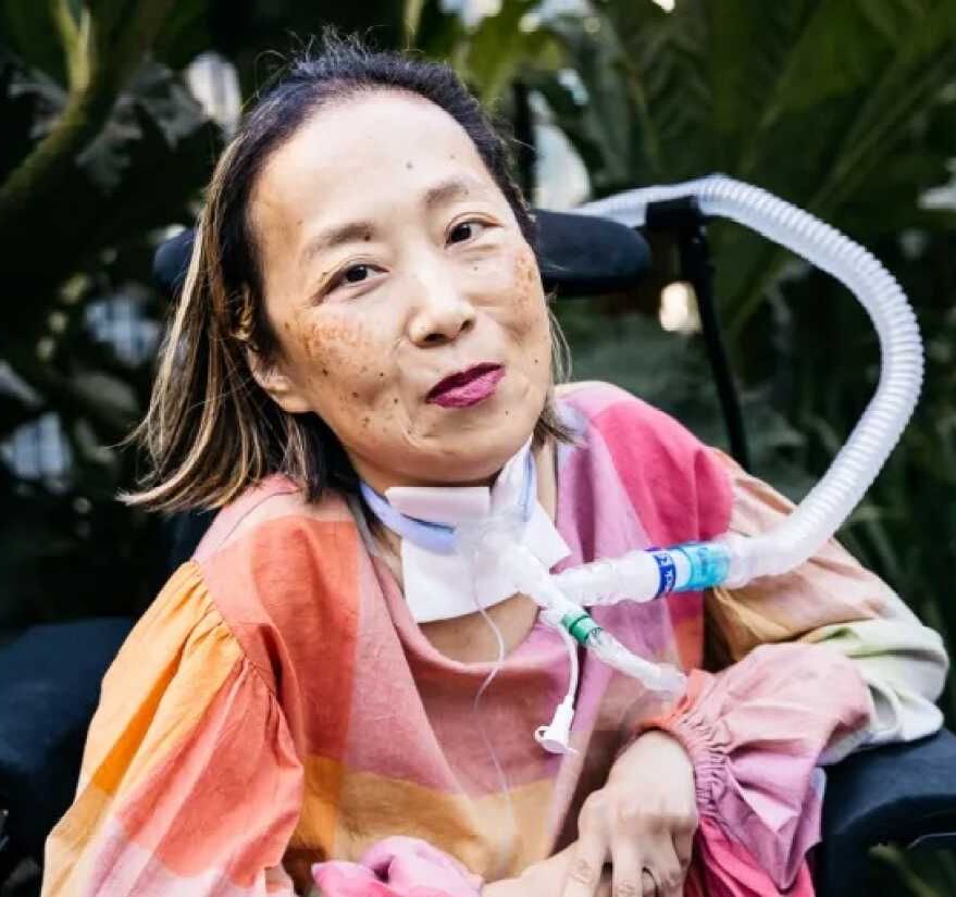 Photo of Alice Wong in headshot style photo taken outside. She is smiling and wearing an orange and pink sweater with greenery visible in background.