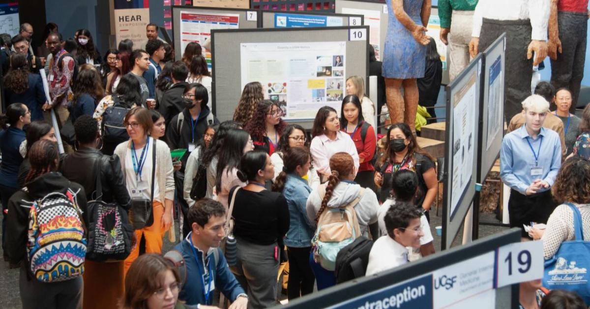 Photo of many people gathered for HEAR Symposium looking at presentations on poster boards in the atrium at Mission Bay.