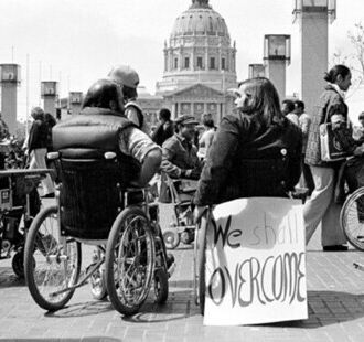 Photo of people who are sitting in wheelchairs protesting in Washington DC. One person has a sign that reads We Shall Overcome in handwritten letters.The White House is seen in the background.  