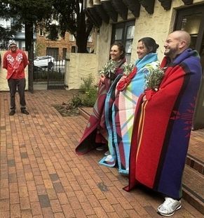 Image of honorees at past Blanket Ceremony, standing with colorful blankets around shoulders and person looking on in background. All are smiling.