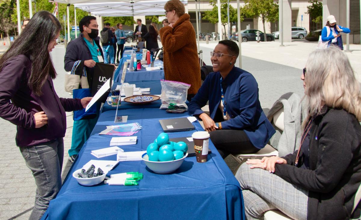 Kendra Hypolite smiles while sitting at table with Denise Caramagno. A person looks at material at a table for Staff Resource Days 2023. They are seated outside with sun shining.
