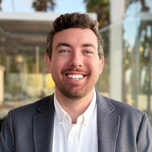 Photo of Asst Director of Jewish Life Daniel Farber smiling in white shirt and grey blazer in headshot style photo outside. 