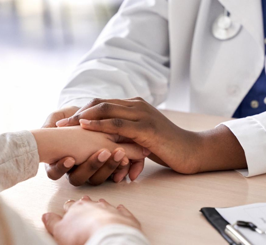 Photo of close-up of clinician in white coat with brown-tone skin holding the hands of a person in their hands. The image is well-lit and supportive feeling.