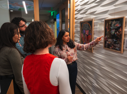 Photo of person showing art work on wall while other people look and listen.