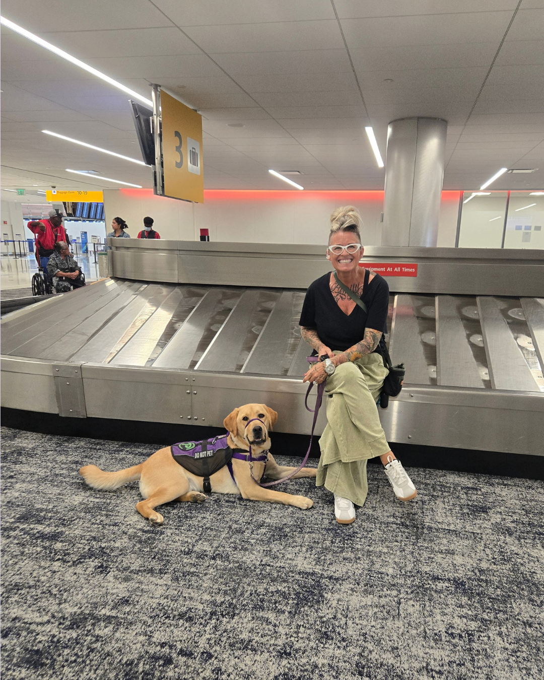 Melissa and Fortune, a yellow lab, pose together near the luggage carousel at the airport. 