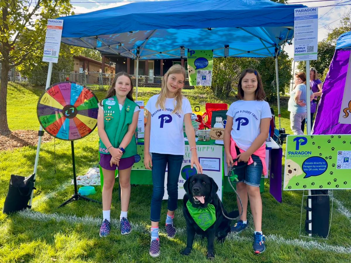 Three girls and a black lab stand in front of a booth featuring a prize wheel and handmade signs inviting people to learn more about Paragon puppy raising.