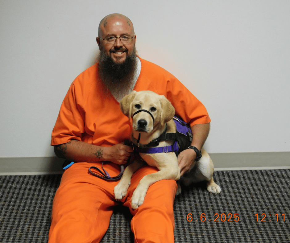 Puppy Fortune lays in his inmate handler, Chris. Chris smiles at the camera. 