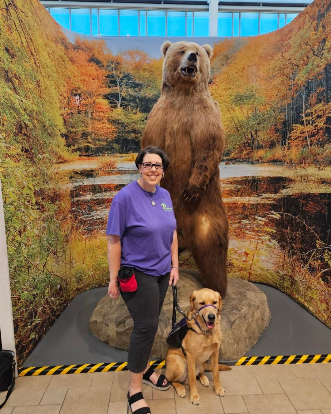 Granby, a Golden/Lab, poses with a volunteer in front of a large brown bear statue.