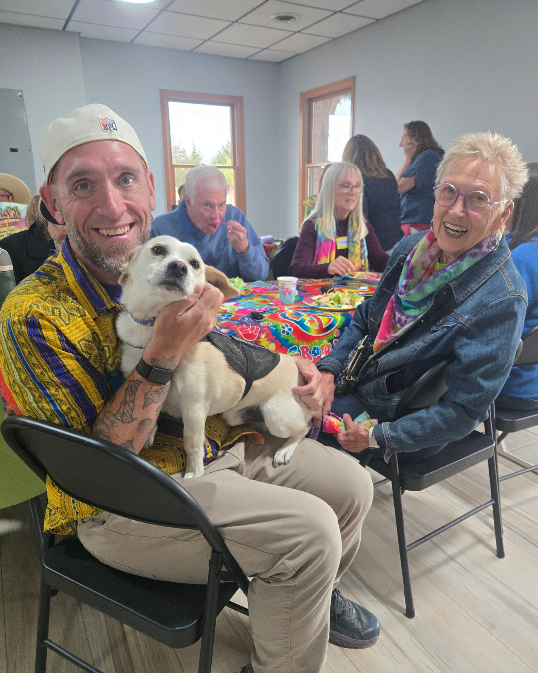 Jack smiles while holding a small white dog named Artie. Arties person, Sue sits next to them smiling. 