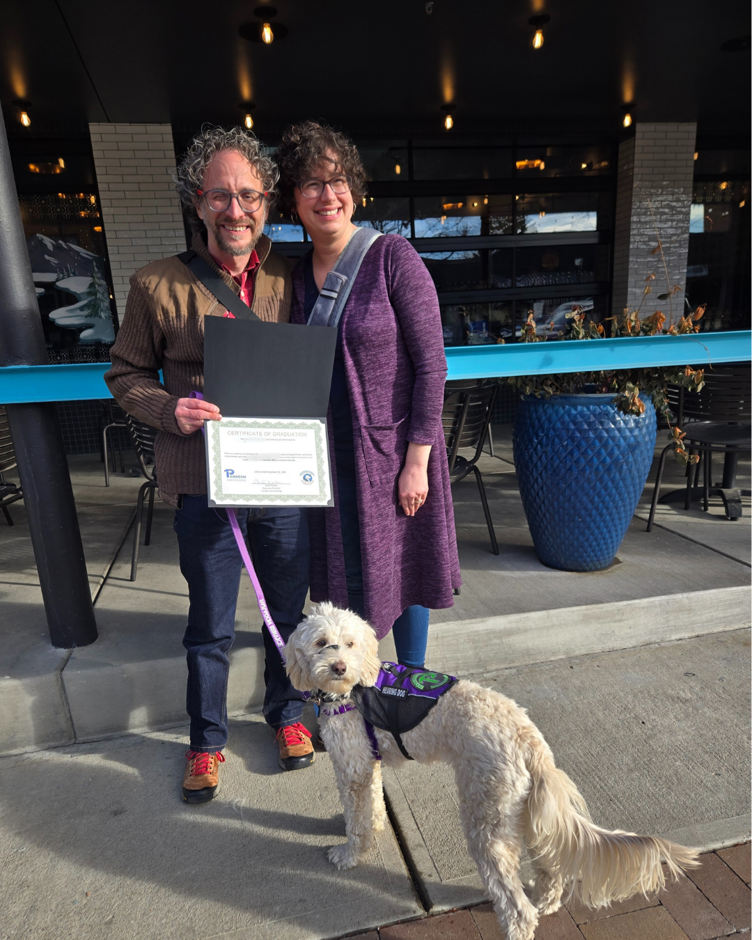 Matt and his wife stand smiling holding a graduation certificate. His hearing dog Eddie stands in front. He's a cream Cobberdog.