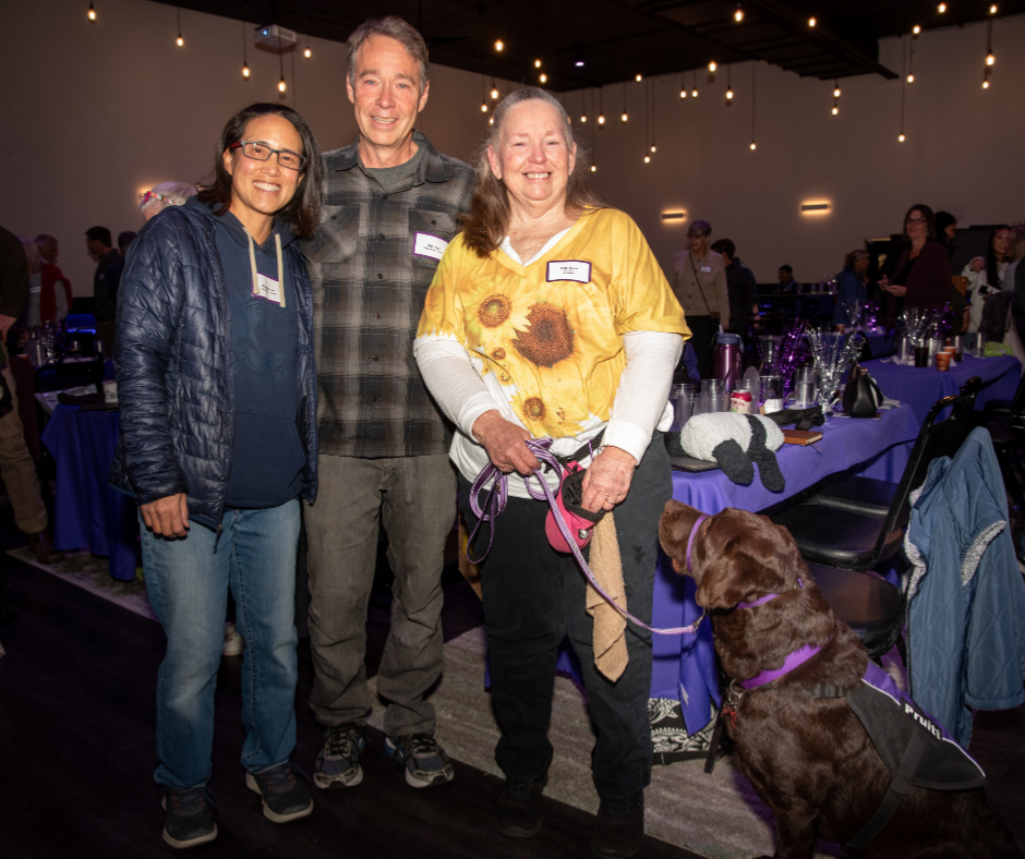 Beth and two others stand smiling at graduation. Pruitt a chocolate lab lays next to her.