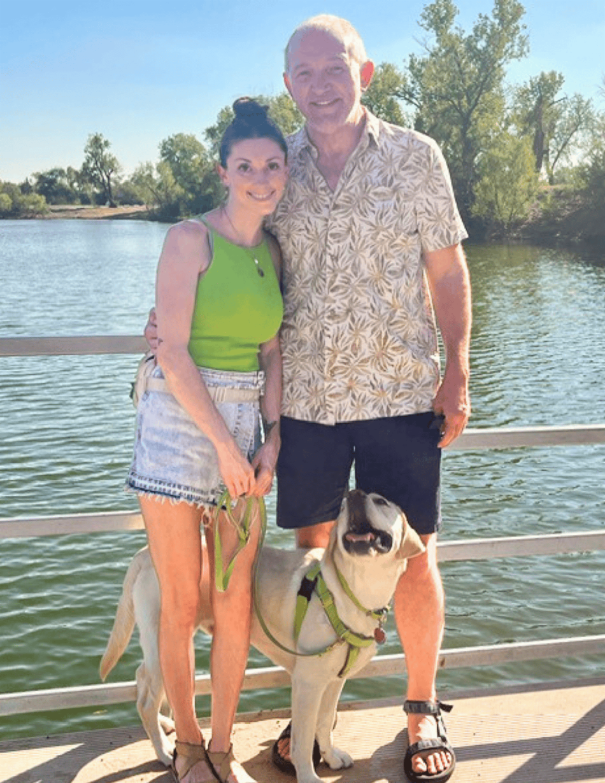 Courtney and her partner Adam stand in front of a lake. A yellow lab named Caro is looking up at them by their feet. 