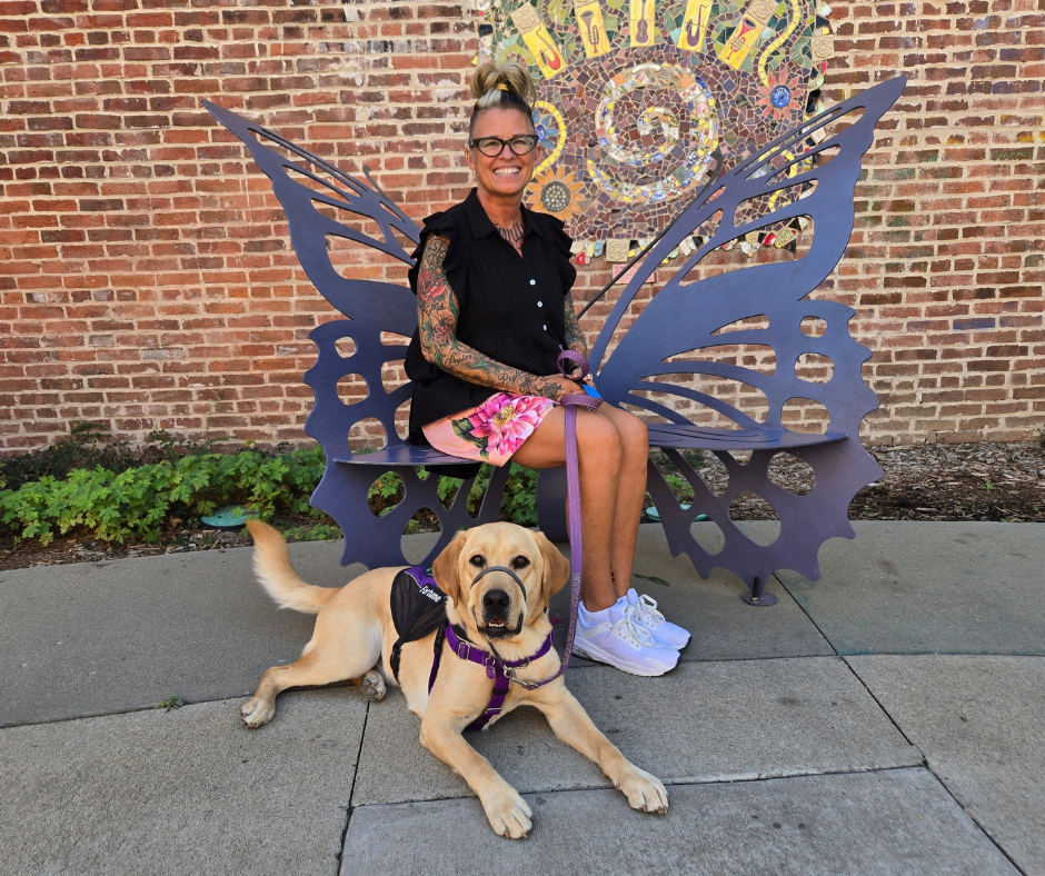 Melissa and Fortune pose on a metal bench in the shape of a butterfly. There is a brick wall in the background. 