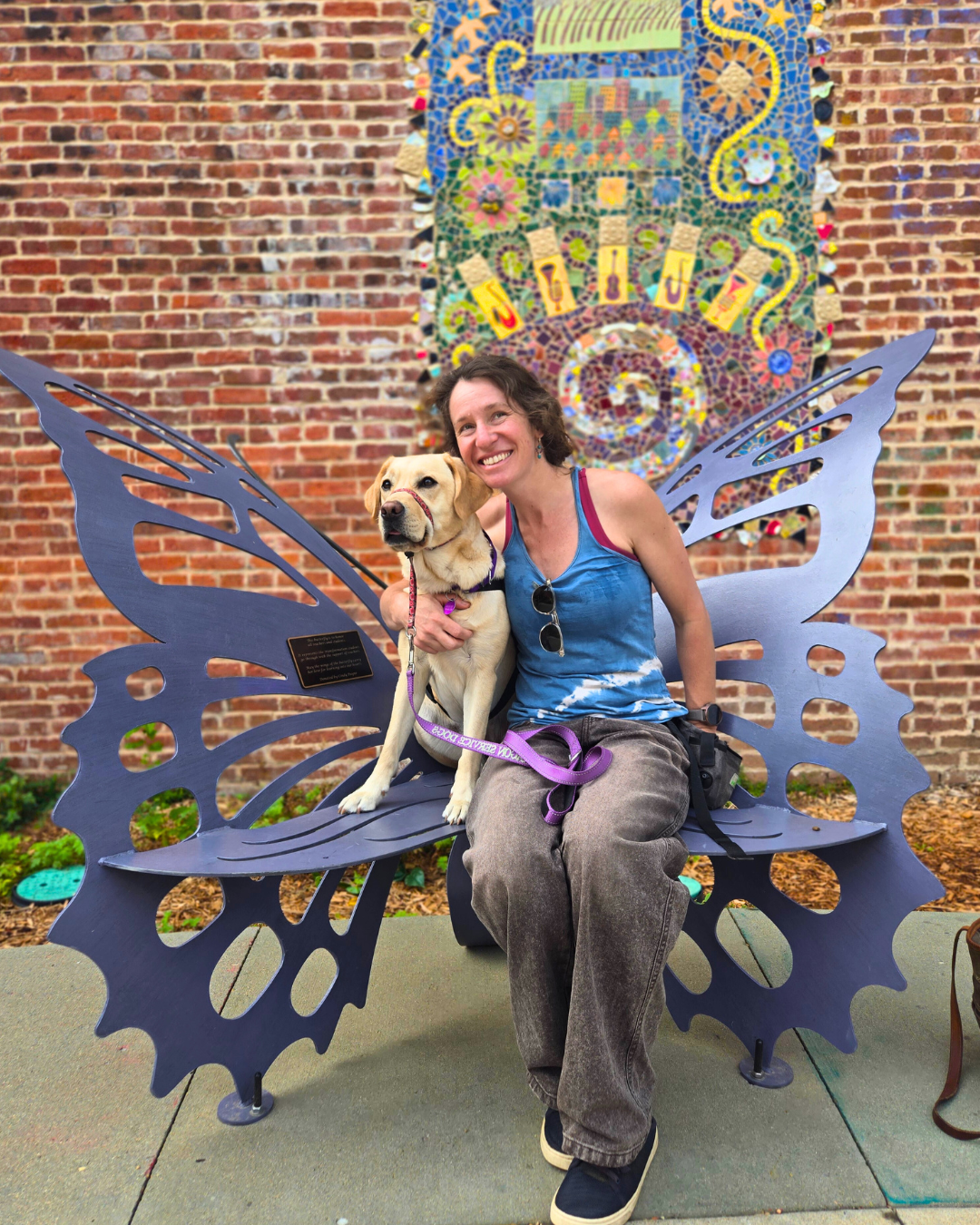 Christina and her hearing dog Danny a yellow Lab sit on a butterfly-shaped metal bench. Behind them a brick wall is painted with a mural of a window and sun. Christina smiles broadly with her arm around Danny.