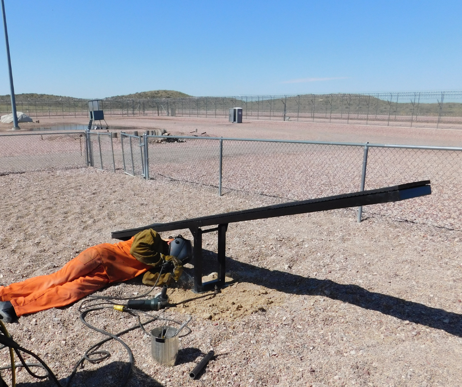 An inmate in a welding mask lays on the ground working under a dog agility teeter. 