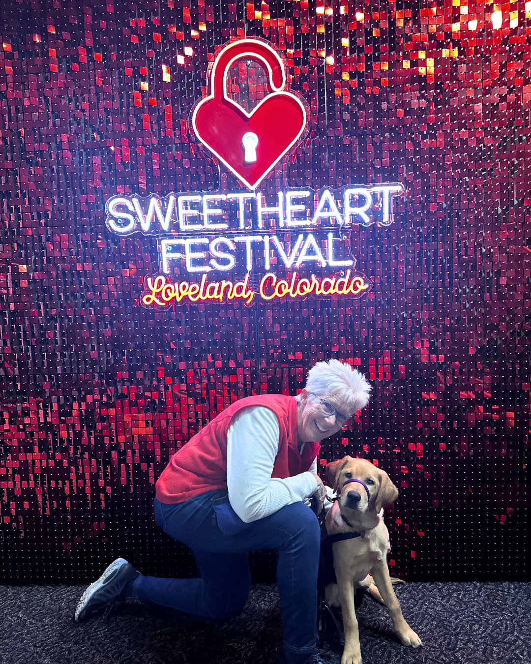 Lori kneels next to a five month old golden lab named Granby. Behind them is a red glowing sign that says "Sweetheart festival Loveland, Colorado".