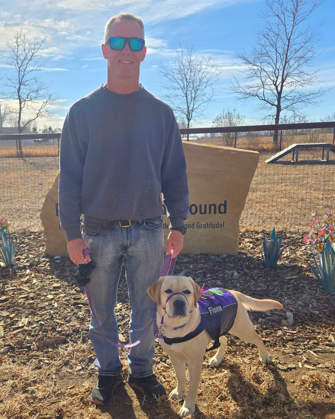 Our volunteer Jim stands next to a yellow lab named Fiona smiling.