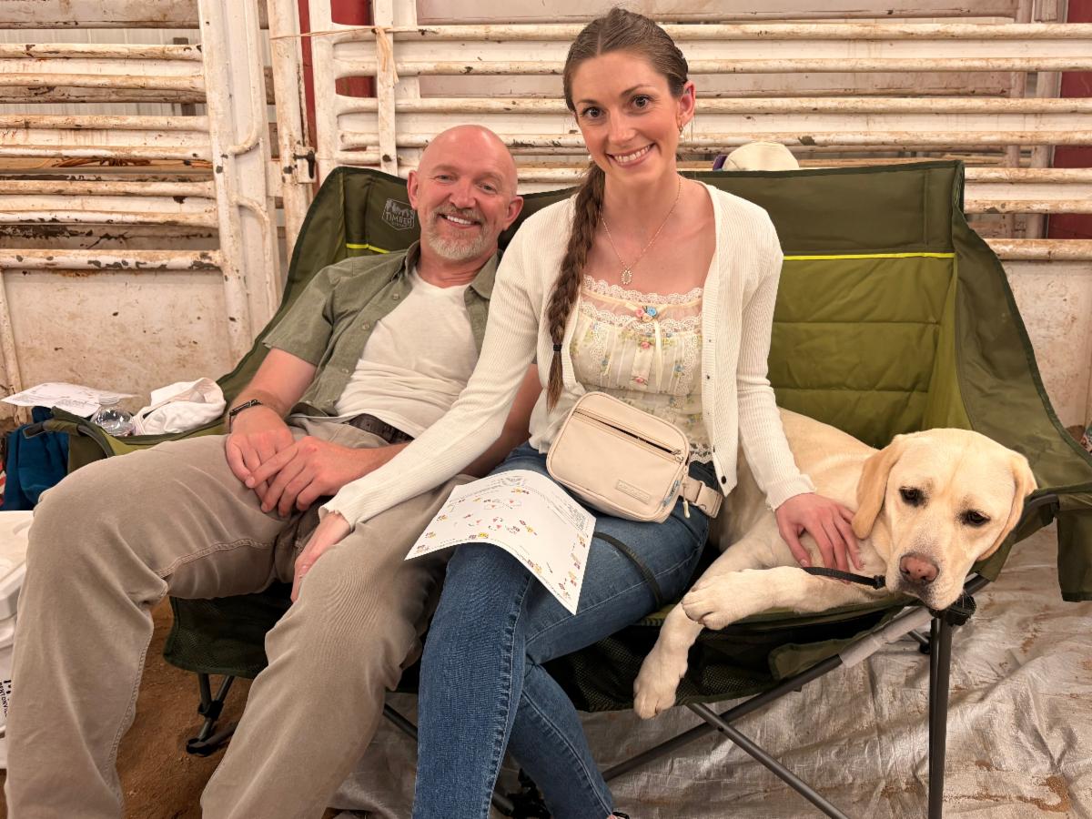 Courtney, Adam, and Caro a yellow lab sit together on a camping chair. 