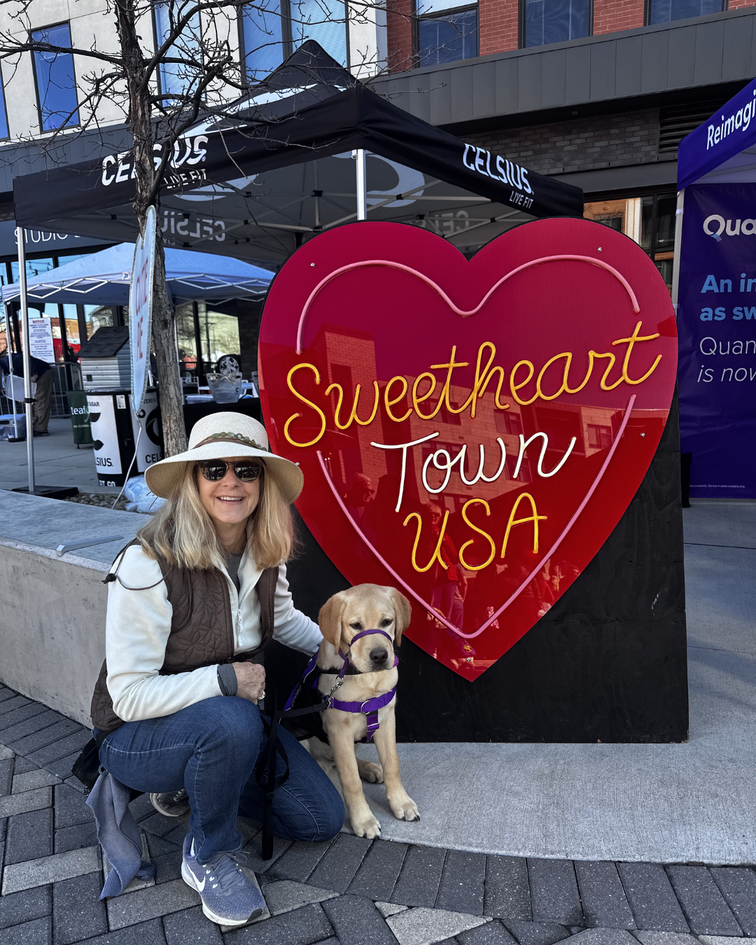 Lisa smiles sitting next to Goose a five month old lab. They are next to a sign that says "Sweet Heart USA". 
