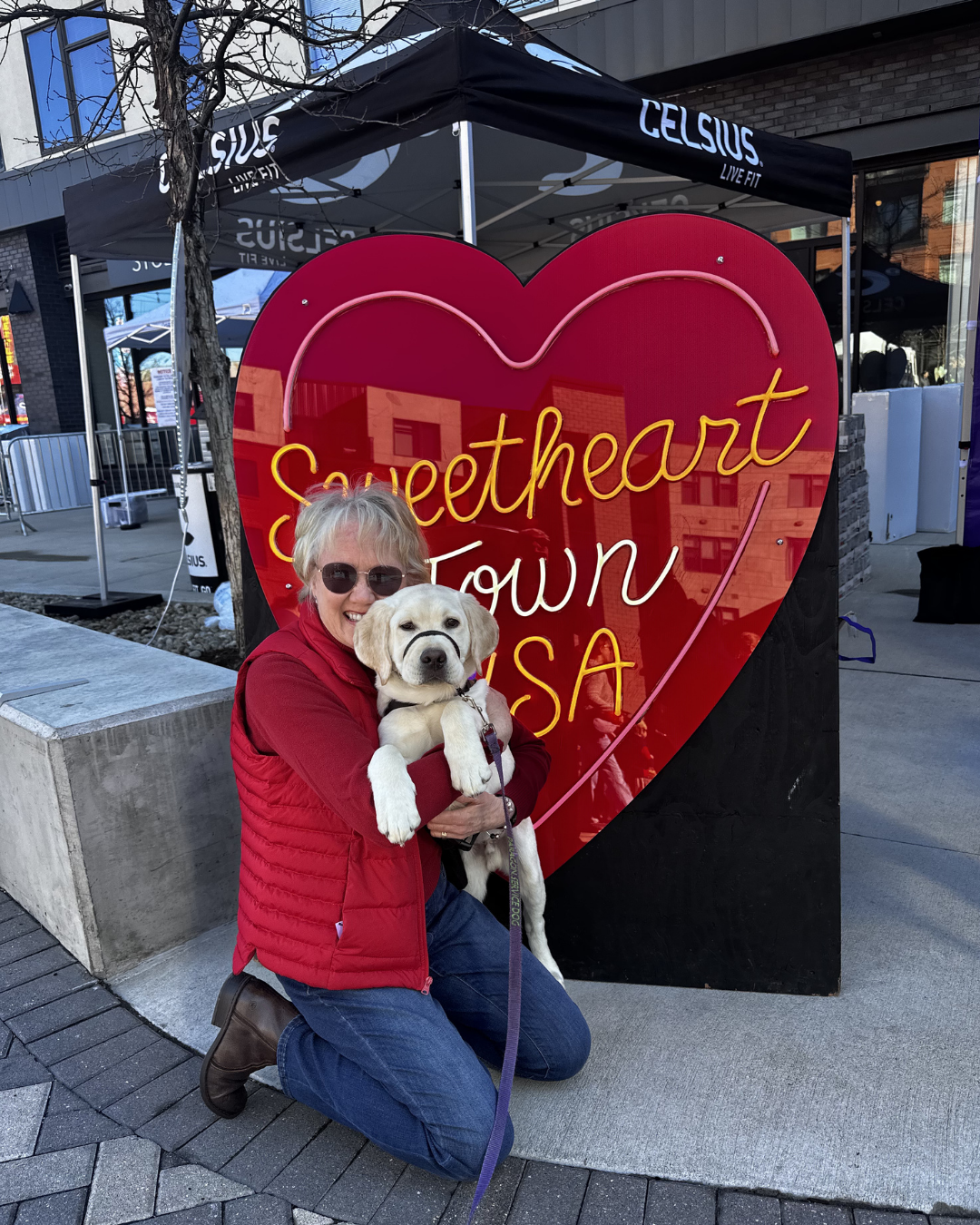 Lisa hold Gilligan a five month old lab. They are in front of a sign that says "Sweet Heart USA". 