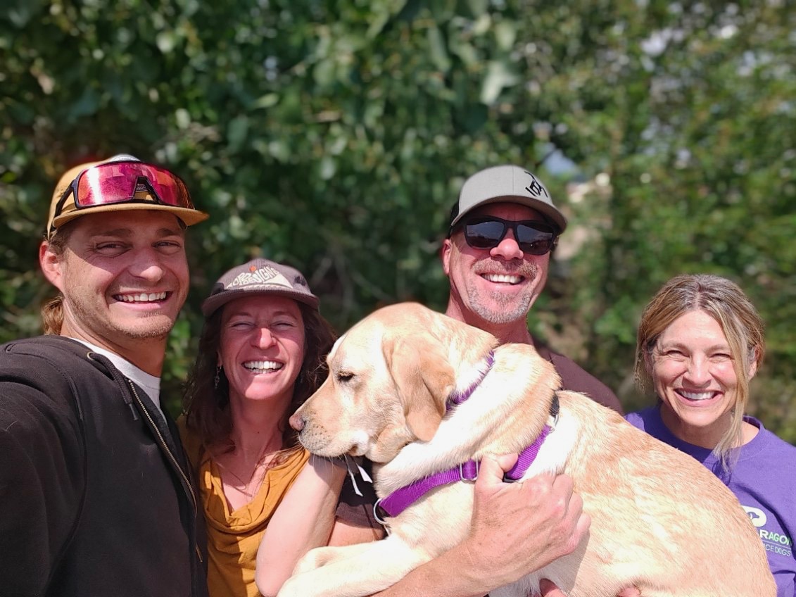Christina and her husband stand with Phil and Heather Dannys puppy raisers. All are smiling at the camera. Phil holds Danny Christinas yellow Lab hearing dog in his arms.