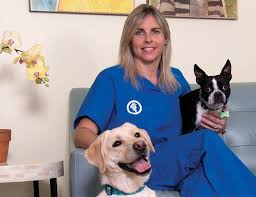 Stacey sits in blue scrubs next to a Frenchie and a yellow lab. 