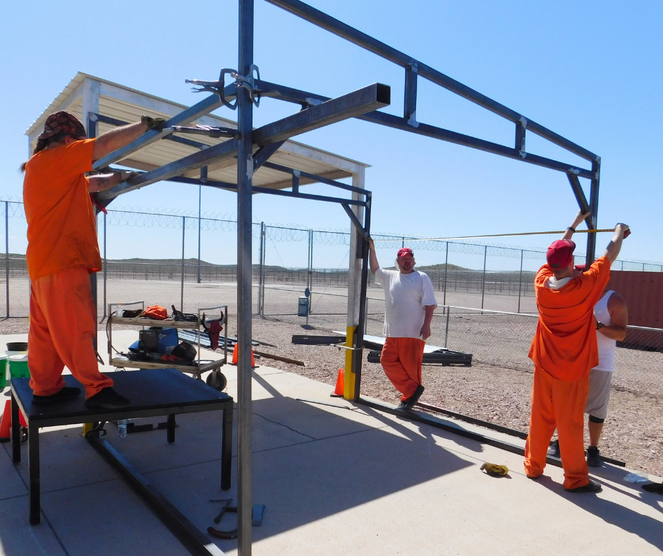 Three inmates hold up various sticks outside as they build a shade structure. 