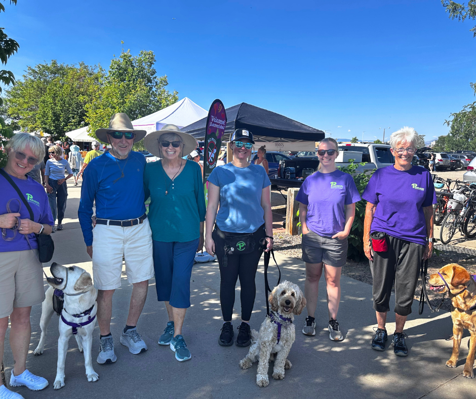 Eight volunteers stand posing at a farmers market. Flapjack, Granby, and Gilligan are at their feet. 