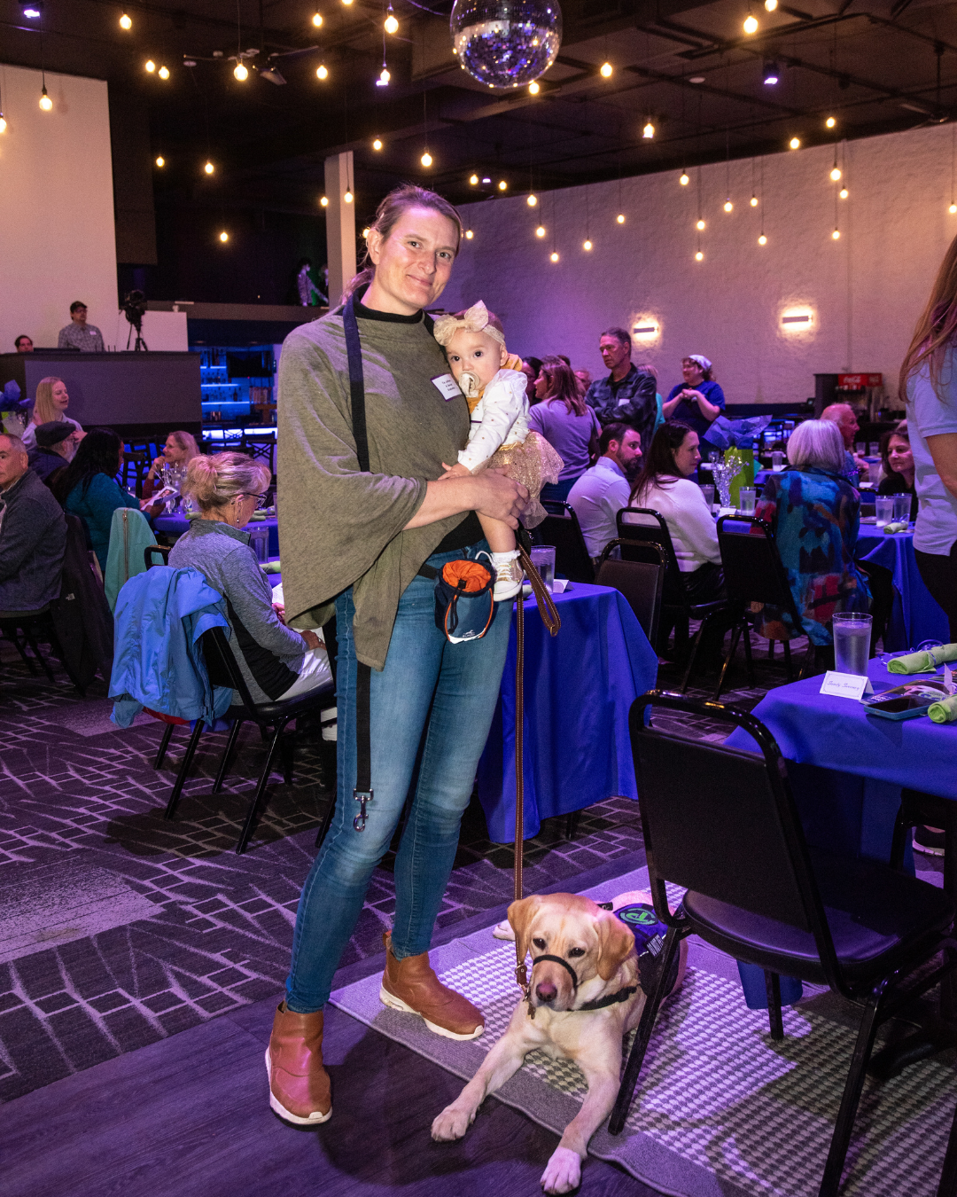 Tia stands holding her six month old baby. Dancer a yellow lab lays at her feet.