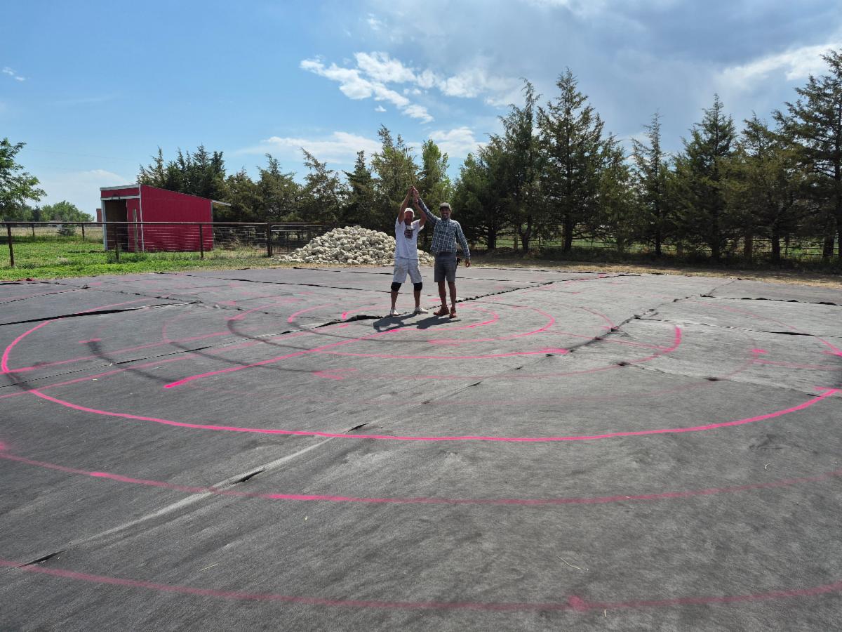 Two men stand in the center of black weed cloth with red lines circular lines. 