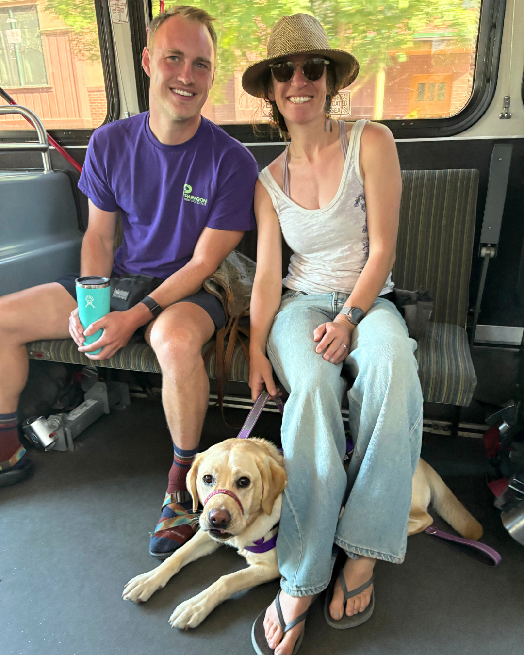 Christina and Paragon trainer Joe sit on a bus together smiling at the camera. Beneath Christinas legs Danny the yellow Lab is tucked in ready for the trip.